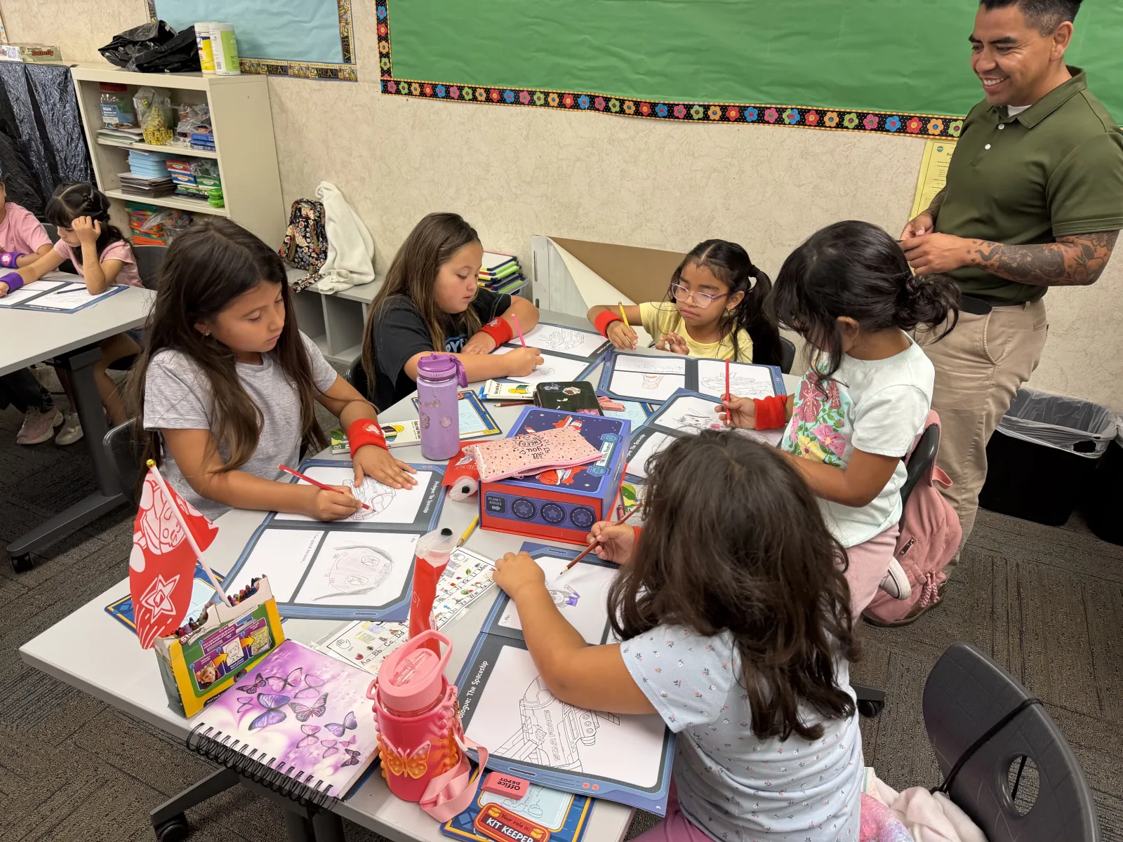 Students color skill worksheets at a table while a facilitator smiles nearby